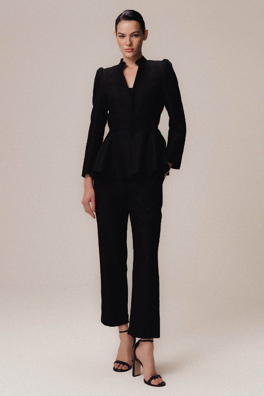 A model wearing a long sleeve black blazer with well-defined lapels and a mandarin collar, paired with matching trousers, standing against a white background.