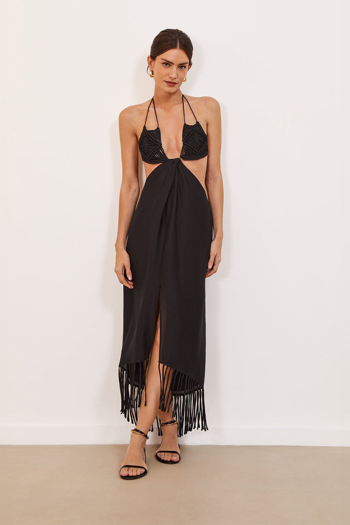 A model wearing a midi black dress with macramé bust, neck and back ties, flowing skirt with fringes, and a front slit.