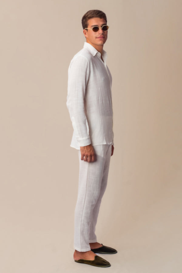 A man standing side-on wearing all white clothes, consisting of a shirt and trousers, with a neutral background.